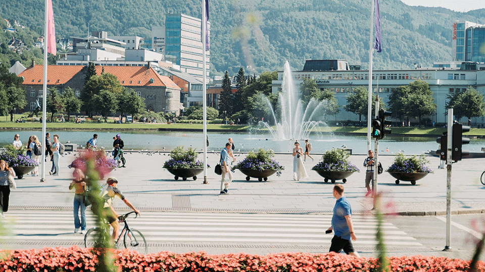 Image of the "Festplassen" in Bergen Centre. In the picture there are people walking and cycling across the square and the crosswalk. The site is paved with stone, with a water in the background. In the water there is a water fountain that creates movement in the image.