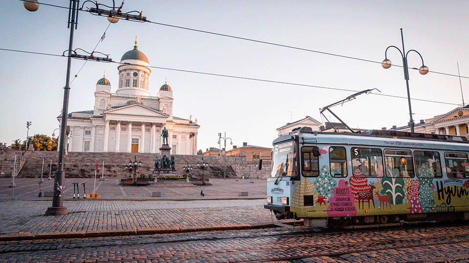 Image of a colorful tram running across a cobbled square. In the background is a tall white, majestic building with statues in front.
