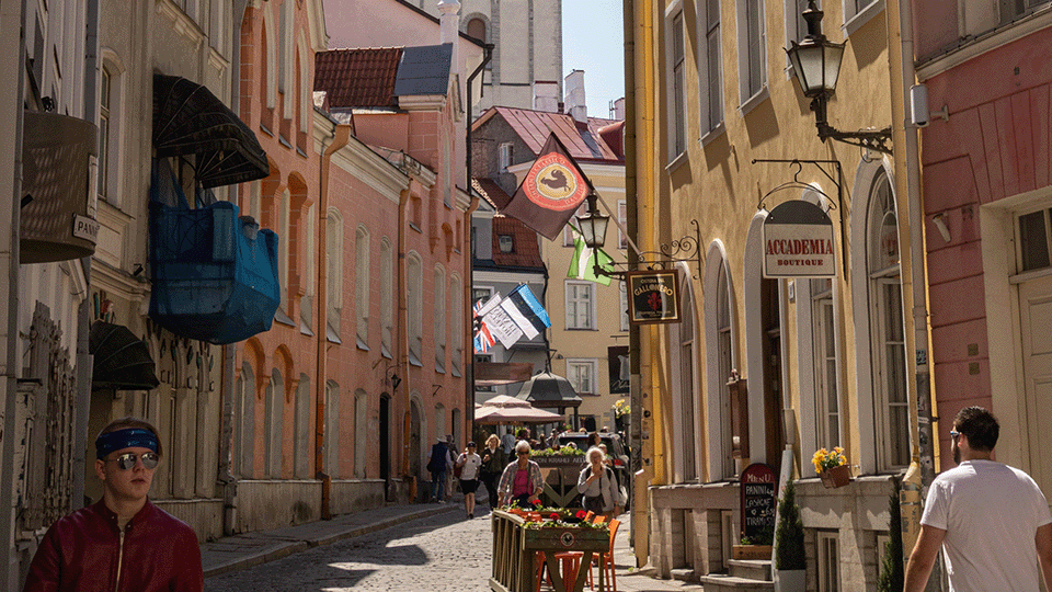 Image of a street between tall buildings in yellow, pink and light green. There are people walking in the street in light summer clothes. The sun shines between the buildings onto the cobblestones.